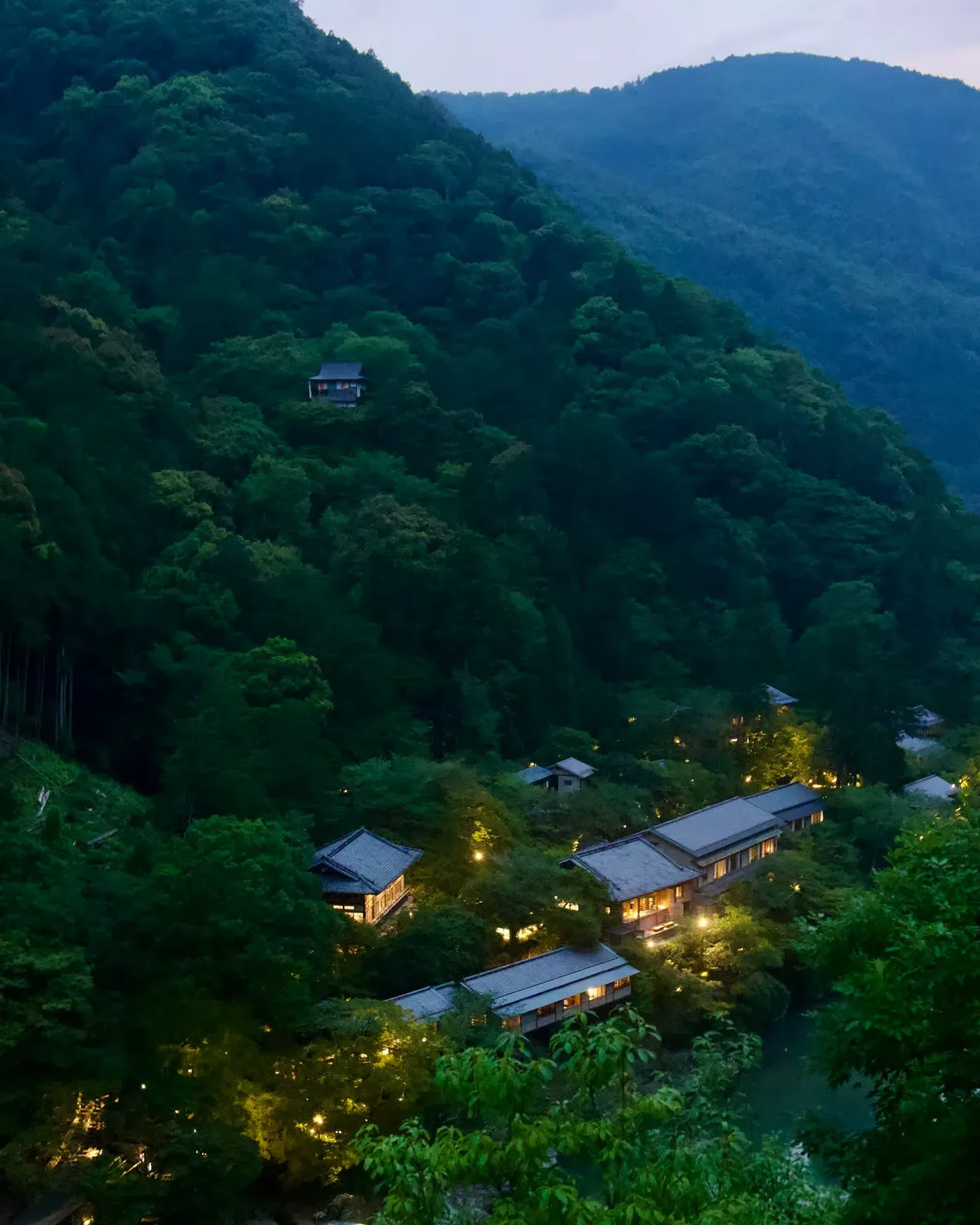 A wooded hillside at dusk above lit inn buildings along the Katsura river, a small temple cabin perched high on the slope