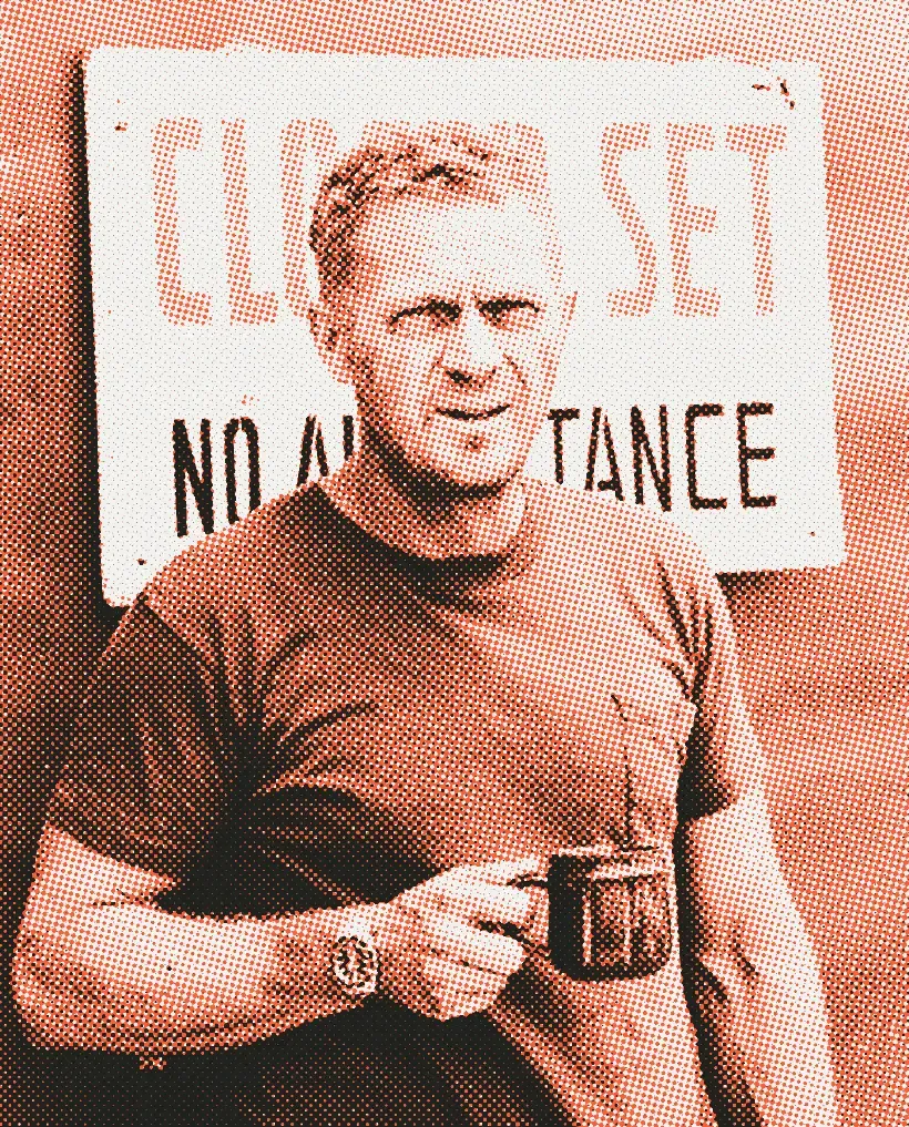 Steve McQueen in a pocket T-shirt holding a mug in front of a “Closed set” sign, a steel diver’s watch on his wrist; black and white