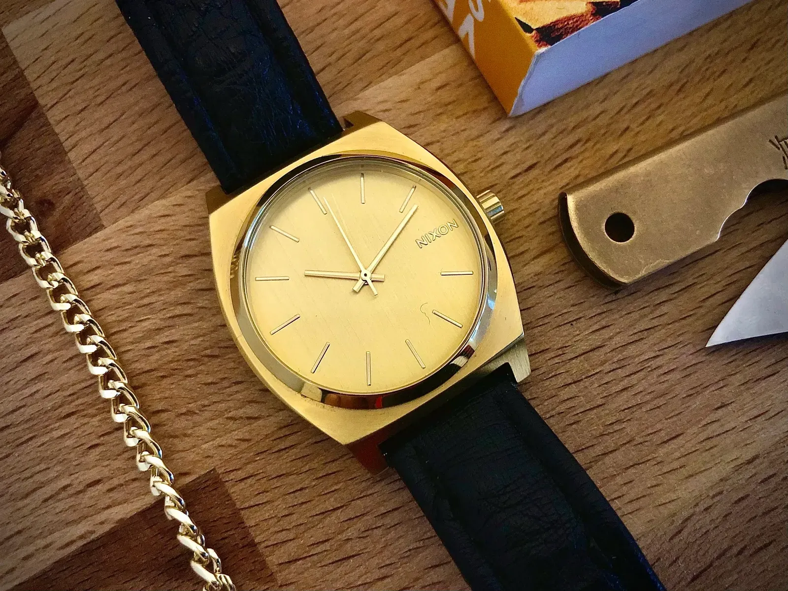 The gold Nixon Time Teller on a black leather strap, lying on a wooden board between a gold chain and a folding knife